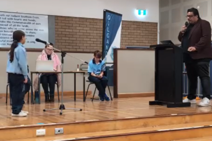 4 People on stage, 2 with microphones for a spelling bee. 2 people are in the background adjudicating the competition.