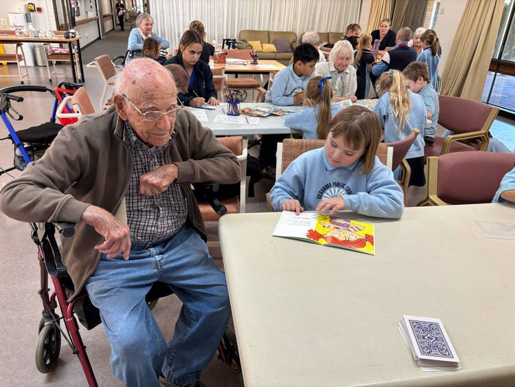 Students visit the residents of the retirement hostel. Foreground student reading to one of the residents. Background students interacting with residents