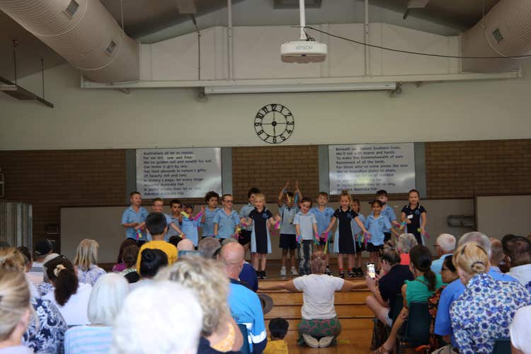 Students on stage in the hall performing. Classroom teacher kneeling, prompting students. Audience in the foreground.